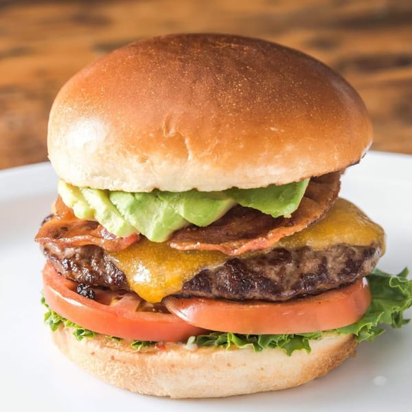 A Sizzlin City Burger with avocado, bacon, melted cheese, tomato, and lettuce on a white plate.
