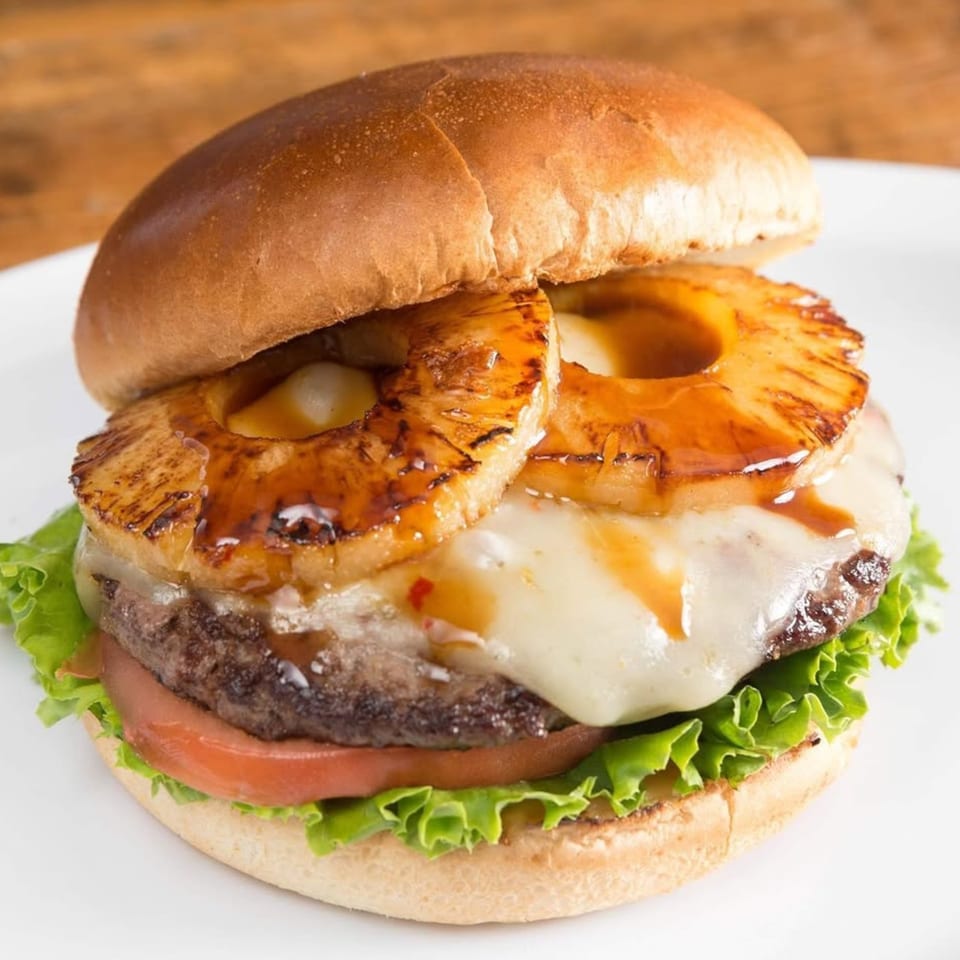 A Sizzlin City Burger with grilled pineapple, melted cheese, lettuce, and tomato on a bun.