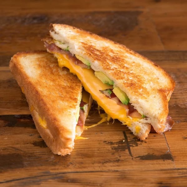 A grilled cheese sandwich with melted cheese, bacon, and avocado, cut in half on a wooden surface.