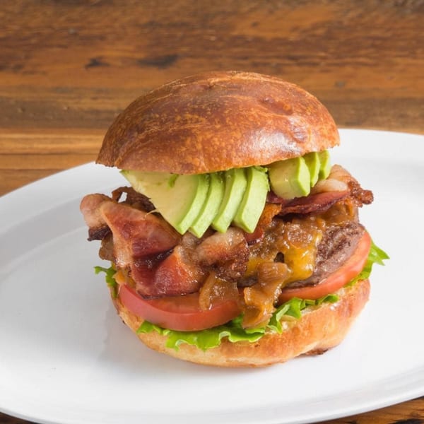 Sizzlin City Burger with bacon, avocado, caramelized onions, tomato, and lettuce on a white plate.