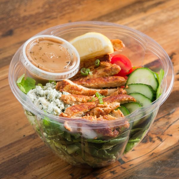 Grilled chicken salad with blue cheese, tomatoes, cucumbers, and dressing in a clear plastic bowl.